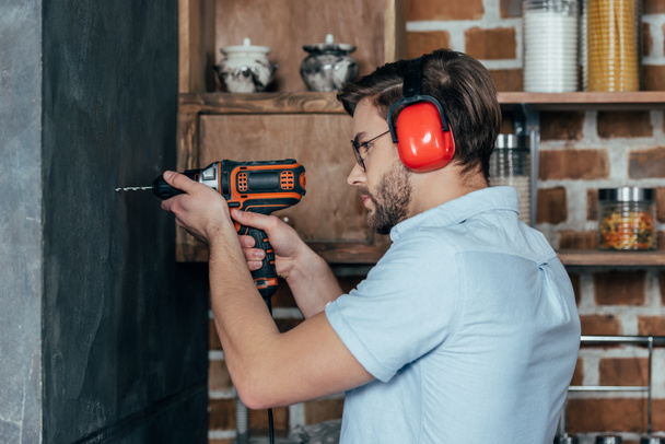 side view of young man in eyeglasses and earmuffs drilling wall with electric drill - Zdjęcie, obraz