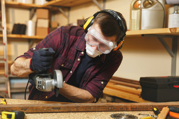 Caucasian young man in plaid shirt, black T-shirt, noise insulated headphones, protective mask working in carpentry workshop at wooden table place with different tools, sawing iron with power saw. - Photo, Image