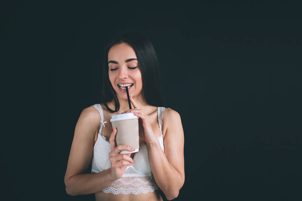 Female model is holding a paper cup of coffee. Young woman with long black hair stands isolated on black background. The girl has a sports figure, she is dressed in white underwear. - Foto, imagen