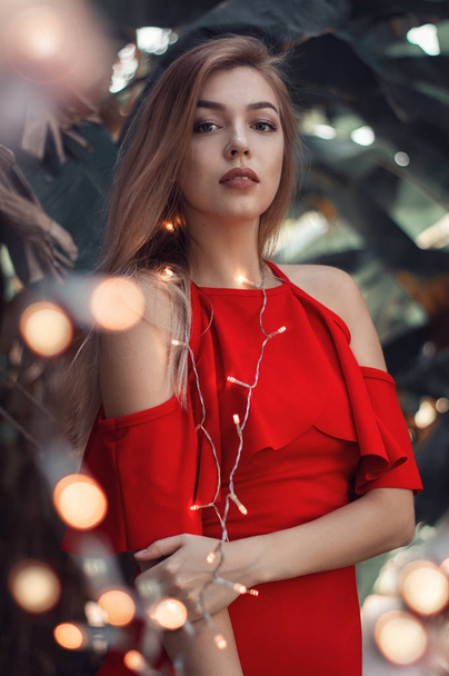 Beautiful young woman with blond hair, in red smart casual dress with small festive lights. Bokeh effect. Portratrait of female model - Фото, изображение