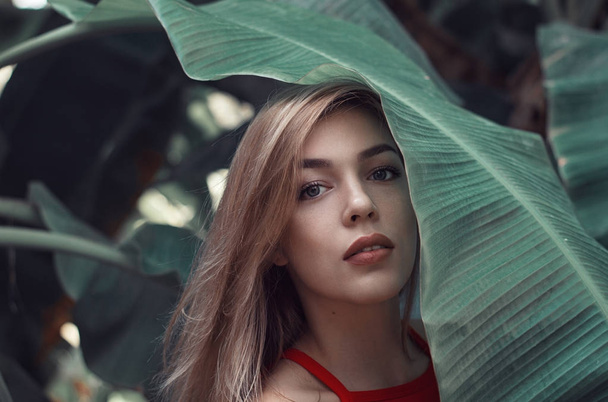 Portrait of a young woman with blond hair standing under a big banana leaf and looking at camera - Фото, зображення