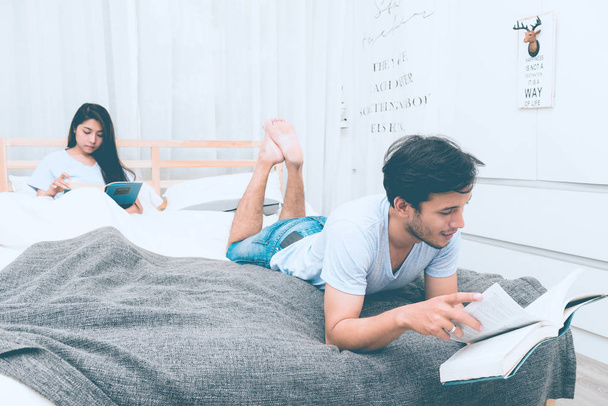 Mixed race lovers concept. Young white male reading a book in his bedroom with his pretty young chinese girlfriend to join. Wearing blue jeans, blue shirt and in their early twenties. Taken indoor. - Fotó, kép