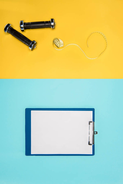 Sports dumbbells, measuring tape and clipboard isolated on yellow and blue - Foto, Bild