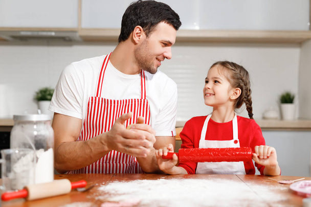 Children, baking, family concept. Cheerful brunet male wears apron and kneads dough, happy girl holds rolling pin, ready to help her father, sit together at kitchen, prepare dough for cookies. - Foto, Bild