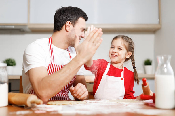 Successful small adorable child and her affectionate father express their agreement between each other, keep hands together, sit at kitchen table, bake delicious apple pie for family on supper. - Foto, Imagem