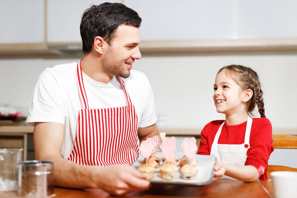 Indoor shot of handsome man wears striped apron and his small pretty female kid hold hand made cakes, rejoices successfully done work on kitchen, being in high spirit, happy spend time together. - Фото, зображення