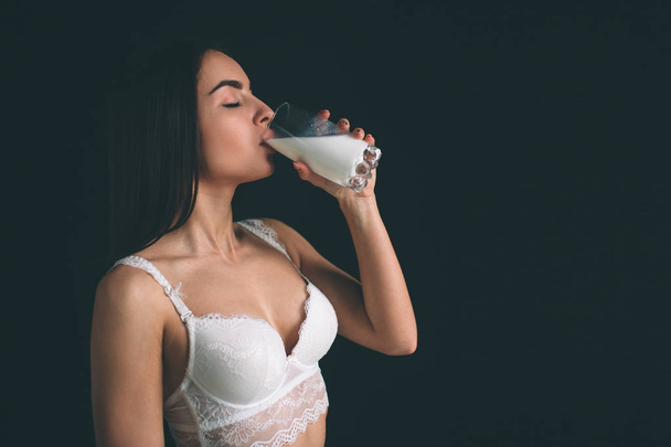Female model drink milk on white background. Young woman with long black hair stands isolated on black background. The girl has a sports figure, she is dressed in white underwear - Fotó, kép