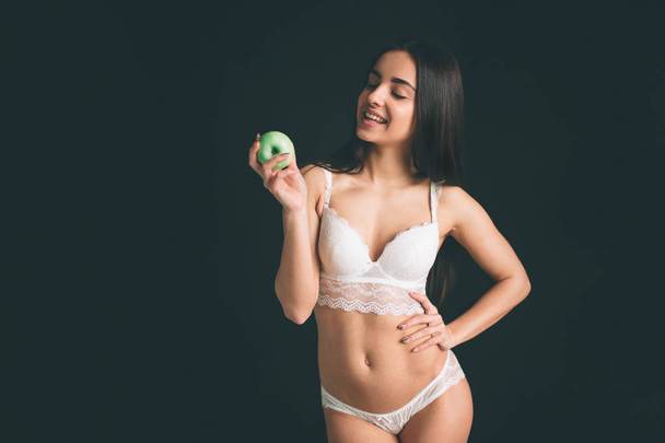 Female model close up with green fresh apple. Vegetarian girl on a diet. Young woman have long black hair isolated on black background. The girl has a sports figure, she dressed in white underwear. - Zdjęcie, obraz