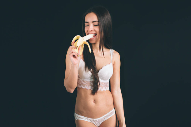 portrait of attractive caucasian smiling woman . studio shot eating banana. Young woman have long black hair isolated on black background. The girl has a sports figure, she dressed in white underwear. - Photo, Image