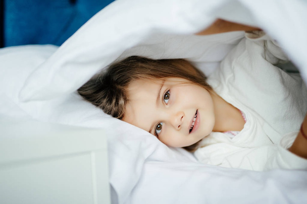 Pretty little girl in sleepwear lying under blanket in bed at home - Fotoğraf, Görsel