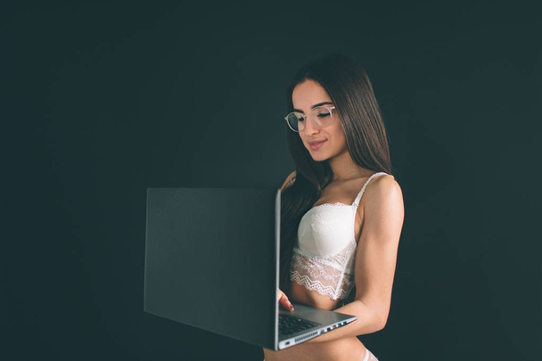 woman use notebook computer. Young woman with long black hair wearing glasses stands isolated on black background. The girl has a sports figure, she is dressed in white underwear - Фото, изображение