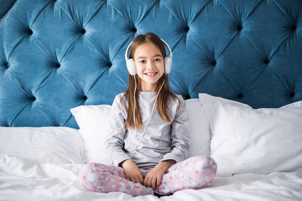Primer plano de linda chica sonriente en los auriculares sentados en la cama, mirando a la cámara en casa
 - Foto, Imagen