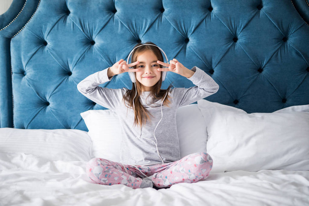 Close-up van schattige lachende meisje hoofdtelefoon zitten in bed, camera thuis kijken - Foto, afbeelding