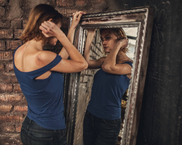 Γυναίκα θύμα του οικιακή βία και κακοποίηση στέκεται κοντά στο καθρέφτη και θέα μώλωπες και πληγές στο πρόσωπο και τα χέρια. - Φωτογραφία, εικόνα