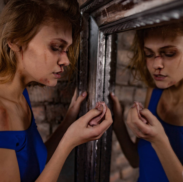 Жертва домашнего насилия стоит возле зеркала с носовым платком и вытирает синяки и раны на лице
. - Фото, изображение