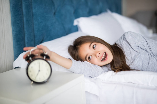 Portrait of shocked cute little girl holding alarm clock and looking at camera while lying in bed in the morning - Fotó, kép