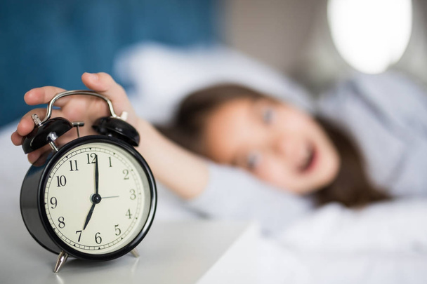 Portrait of shocked cute little girl holding alarm clock and looking at camera while lying in bed in the morning - Photo, Image