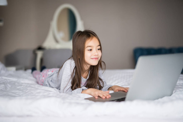 zufriedenes begeistertes kleines Mädchen, das auf dem Bett sitzt, während es lächelnd den Film ansieht, während der Laptop vor ihr platziert wird - Foto, Bild