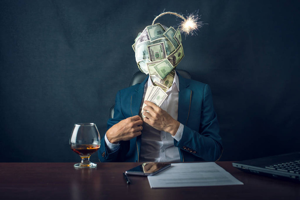 A businessman sitting on the workplace and putting the bribe money in his pocket with a bomb in the form of a ball dollar bills instead of his head. The concept of risk of financial fraud. - Photo, Image