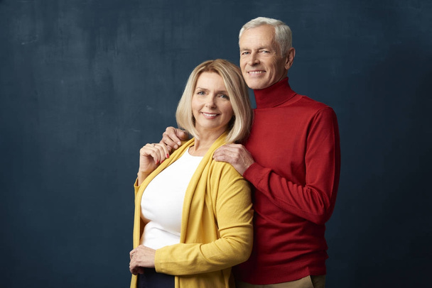 Happy senior couple standing at dark background. Beautiful mature woman looking at camera and smiling while elderly man embracing her. - Фото, изображение