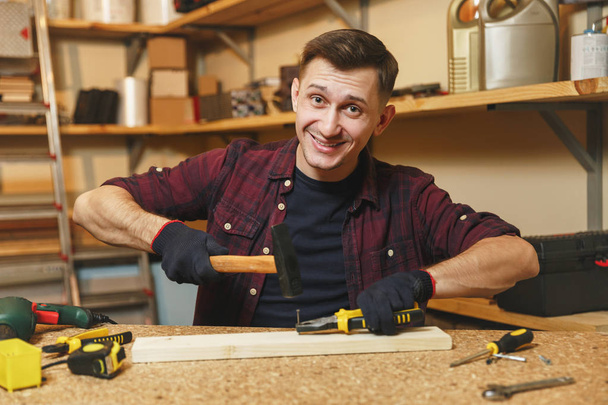 Handsome smiling caucasian young man in plaid shirt, black T-shirt, gloves hammering nails with hammer, working in carpentry workshop at wooden table place with piece of wood, different tools. - Photo, Image