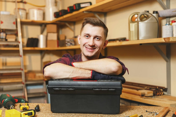 Handsome charismatic caucasian young man in plaid shirt, black T-shirt, gloves working in carpentry workshop at wooden table place, holding hands crossed on black toolbox with different tools. - Photo, Image
