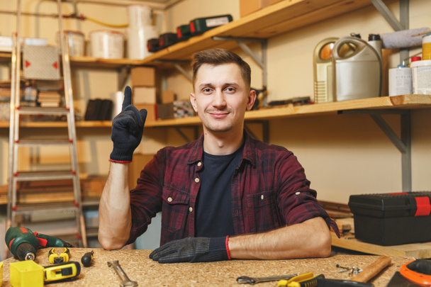 Handsome smiling caucasian young man in plaid shirt, black T-shirt, gloves pointing index finger up, working in carpentry workshop at wooden table place with piece of wood, different tools. - Photo, Image