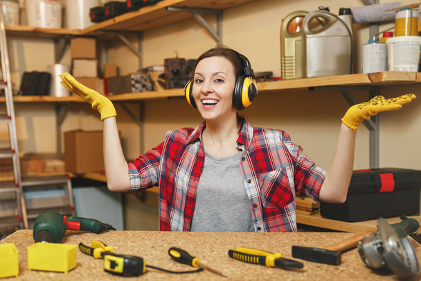 Young woman in plaid shirt, gray T-shirt, noise insulated headphones, yellow gloves spreading hands, working in carpentry workshop at wooden table place with piece of wood, different tools. Copy space - Photo, Image