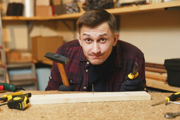 Handsome smiling caucasian young man in plaid shirt, black T-shirt, gloves hammering nails with hammer, working in carpentry workshop at wooden table place with piece of wood, different tools. - 写真・画像