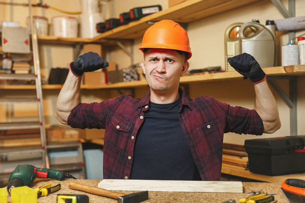 Красивый сильный кавказский юноша в клетчатой рубашке, черная футболка, оранжевый защитный шлем, перчатки, работающие в столярной мастерской на деревянном столе с куском дерева, молотком, различными инструментами
. - Фото, изображение