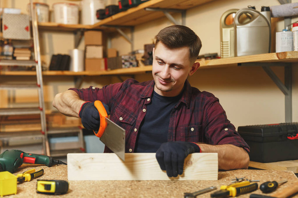 Handsome charismatic smiling caucasian young man in plaid shirt, black T-shirt, gloves sawing wood with saw, working in carpentry workshop at wooden table place with piece of wood, different tools. - Photo, Image