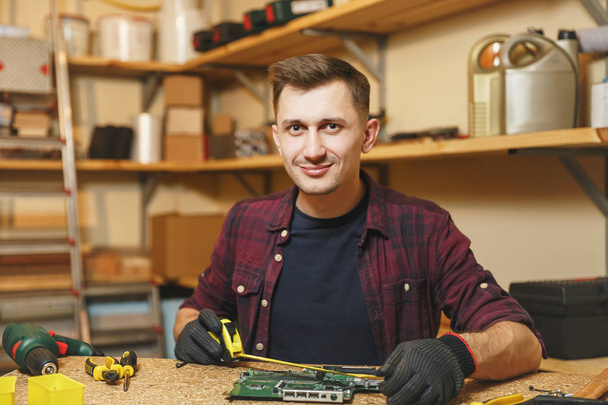 Multiméter a villanyszerelő. Kaukázusi fiatalember, kockás ing, fekete póló digitális elektronikus mérnök javítása, forrasztási számítógép Pc alaplap workshop-on fából készült asztal különböző eszközök. - Fotó, kép