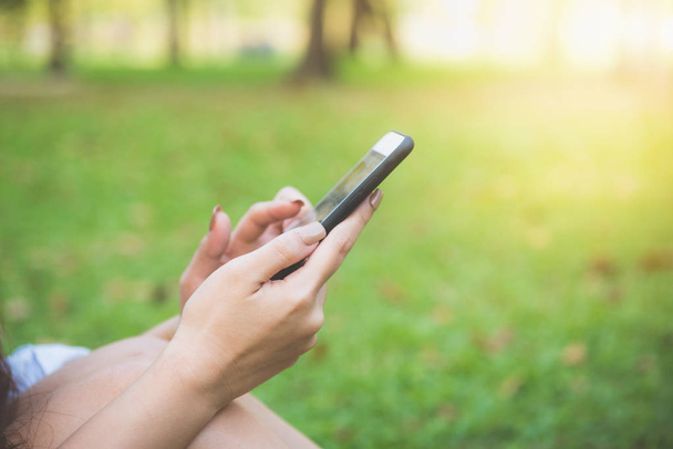 Söpö aasialainen nainen lukee miellyttävää tekstiviestiä matkapuhelimella istuessaan puistossa kevätpäivänä. Aasian nainen käyttää älypuhelimella tunne rentoutua ja hymynaama. Elintapojen ja teknologian käsitteet
. - Valokuva, kuva