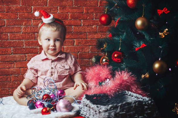 Красива дитина в капелюсі Санта сидить біля ялинки з різдвяними іграшками і вогнями
 - Фото, зображення
