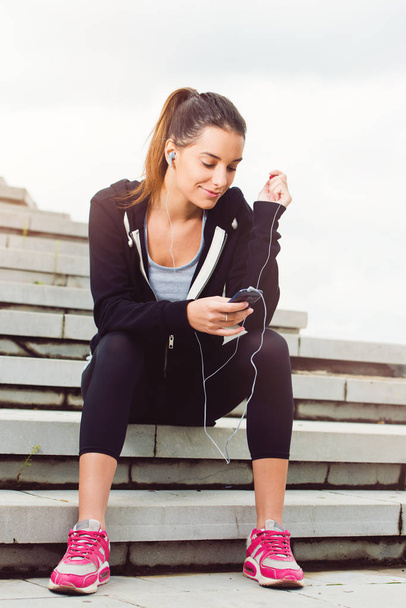 Молодая женщина отдыхает от тренировок на улице с сотовым телефоном
 - Фото, изображение