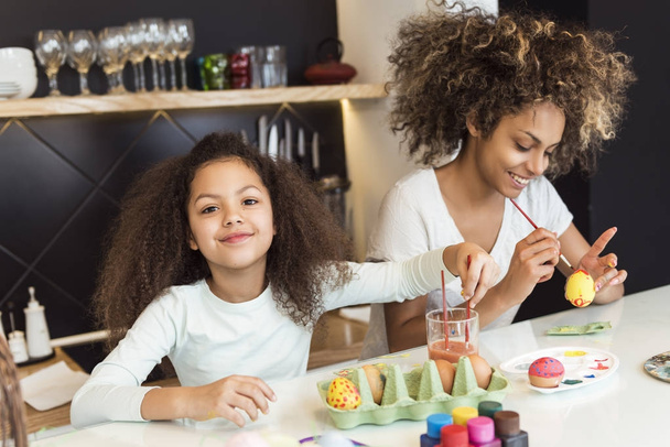 Beautiful African American woman and her daughter coloring Easter eggs in the kitchen - Zdjęcie, obraz