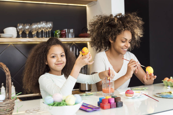 Beautiful African American woman and her daughter coloring Easter eggs in the kitchen - Zdjęcie, obraz