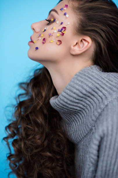 side view of young woman with flowers on face in warm sweater isolated on blue - Foto, imagen