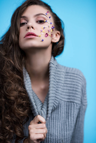 young woman in stylish turtleneck sweater with flowers on face isolated on blue - Fotó, kép