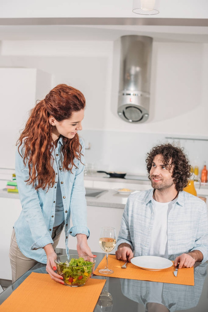 smiling girlfriend putting salad on table and looking at boyfriend - 写真・画像