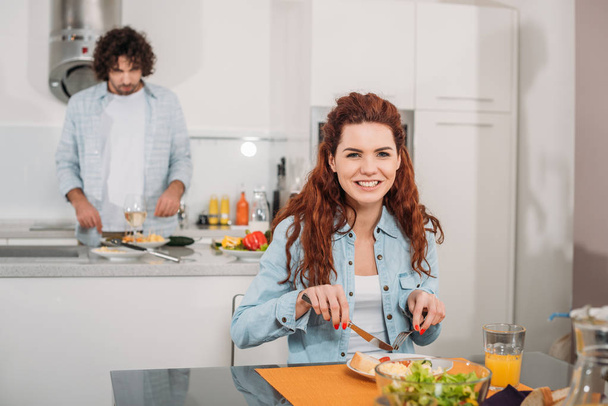 smiling girlfriend eating while boyfriend cooking at kitchen - Photo, Image