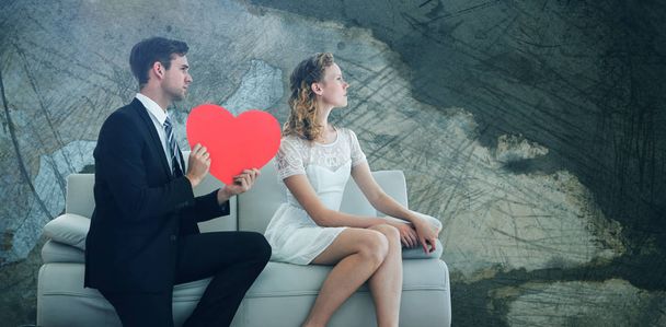 Man showing red heart shaped paper to woman while sitting on sofa  against rusty weathered wall - Photo, image