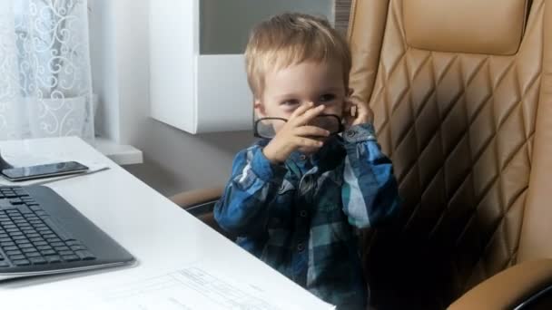 4k footage of adorable smiling toddler boy sitting in big boss chair at office and wearing eyeglasses. Concept of little businessman - Záběry, video