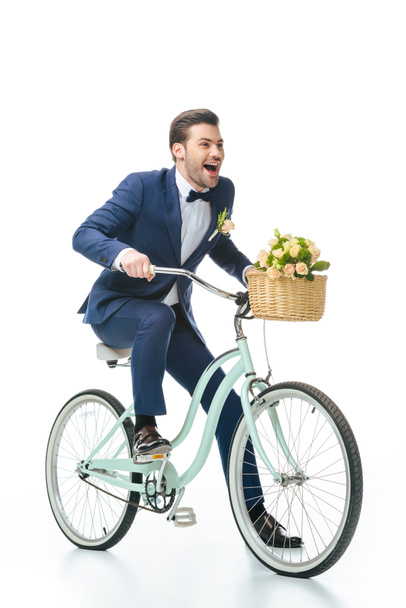 happy groom in suit riding retro bicycle with wedding bouquet in basket isolated on white - Foto, imagen