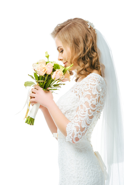 Seitenansicht der attraktiven Braut in Brautkleid und Schleier mit Blumenstrauß isoliert auf weiß - Foto, Bild