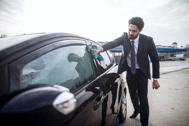 handsome stylish young bearded man in suit cleaning car with microfiber cloth at the manual car washing self-service station - Foto, immagini
