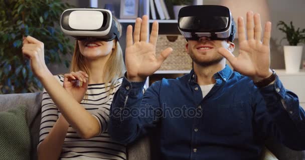 Close up of the young couple wearing VR glasses sitting on the couch in the living room and having VR headset with many emotions, waving their hands in the air. Evening time. Portrait. Indoor - Imágenes, Vídeo