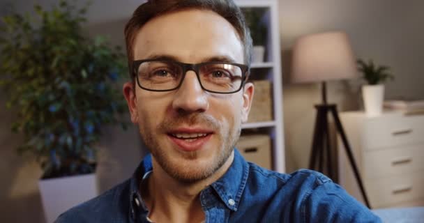 Young Caucasian man in glasses videochatting, waving his hand and giving a thumb up while sitting in the living room in the evening. POV. Portrait. Close up. Indoors - Кадры, видео