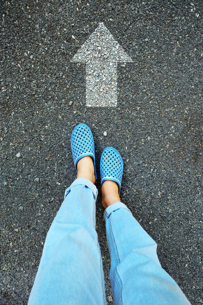Selfie μπλε παπούτσια και μπλε τζιν. Γυναικεία πόδια καθώς στέκεται όρθιος με λευκό βέλος γραμμή στο δρόμο τσιμέντου φόντο μεγάλη για οποιαδήποτε χρήση. - Φωτογραφία, εικόνα