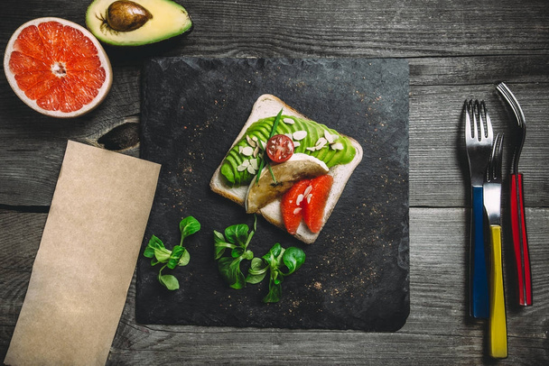 Voeding biologische toast met avocado, grapefruit en amandel schilfers. Leisteen zwart plaat, vorken, houten grijze achtergrond, schone papierruimte, top uitzicht - Foto, afbeelding